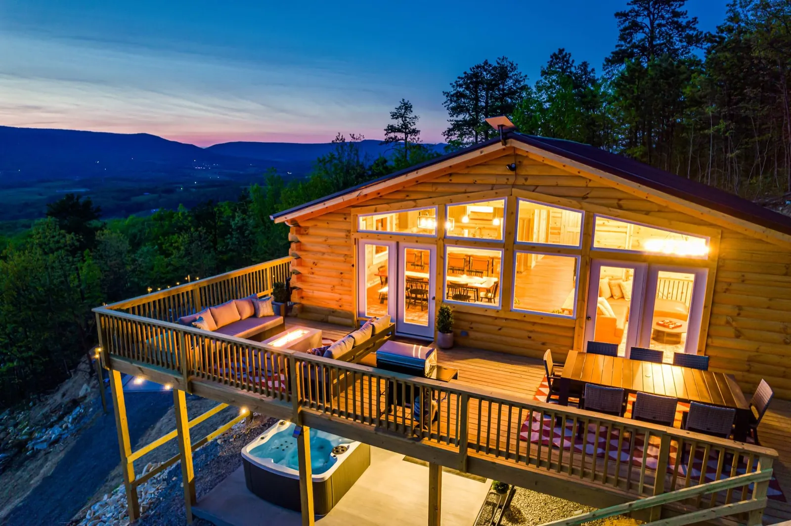 Wildwood Lodge at twilight: the log cabin’s glowing wall of windows above a furnished deck and hot tub, with the Shenandoah Valley behind