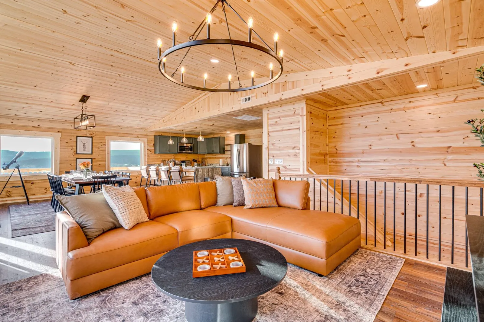 The upstairs of Wildwood Lodge: a cognac leather sectional in front, the long dining table and kitchen beyond, under a vaulted pine ceiling